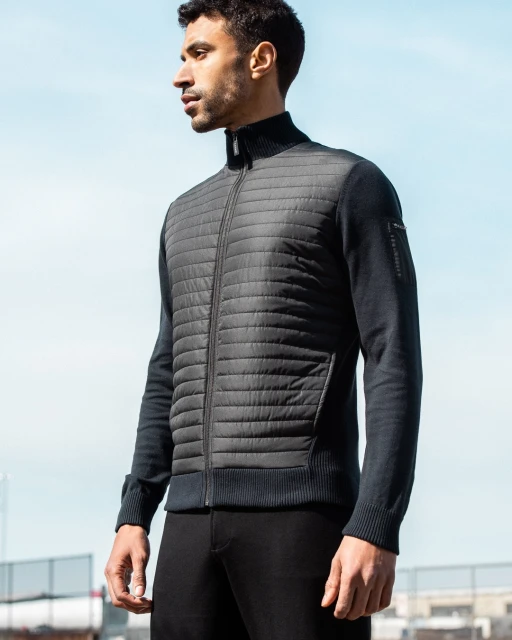 A man stands in a medium shot, wearing a black quilted jacket and black pants, against a light blue sky, showcasing outdoor fashion.
