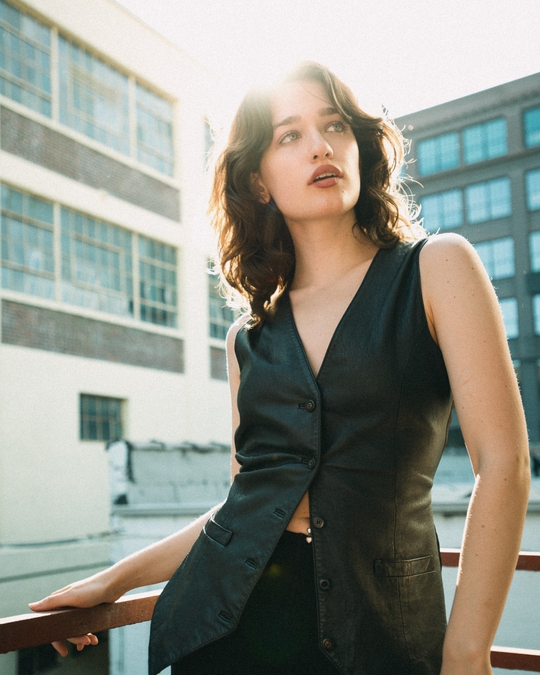 Young woman in a black leather vest leaning against a railing outdoors with brick and glass architecture behind her.