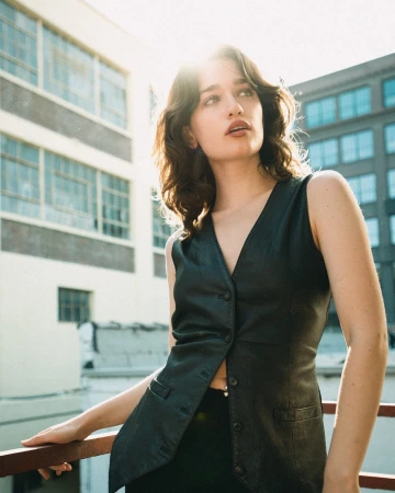 Young woman in a black leather vest leaning against a railing outdoors with brick and glass architecture behind her.