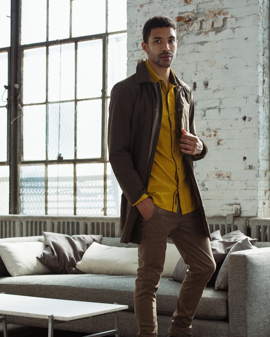 Stylish man with brown hair and coat poses indoors. He wears a yellow shirt and brown pants, standing near a grey couch and window.