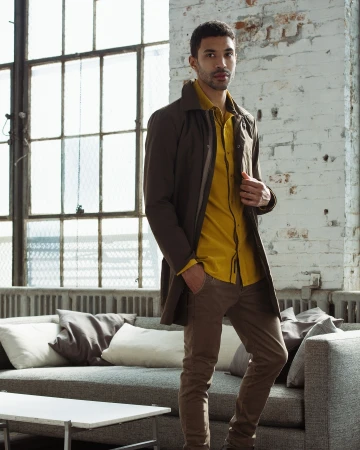 Stylish man with brown hair and coat poses indoors. He wears a yellow shirt and brown pants, standing near a grey couch and window.