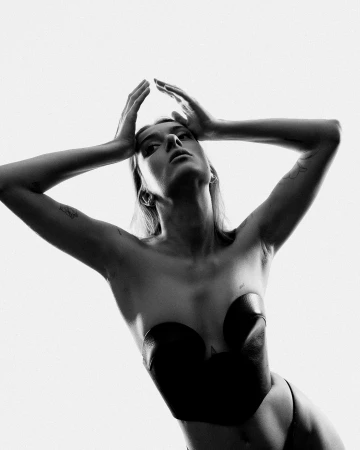 Striking black and white studio portrait of a woman in a structured black top with her arms raised, set against a stark white backdrop.