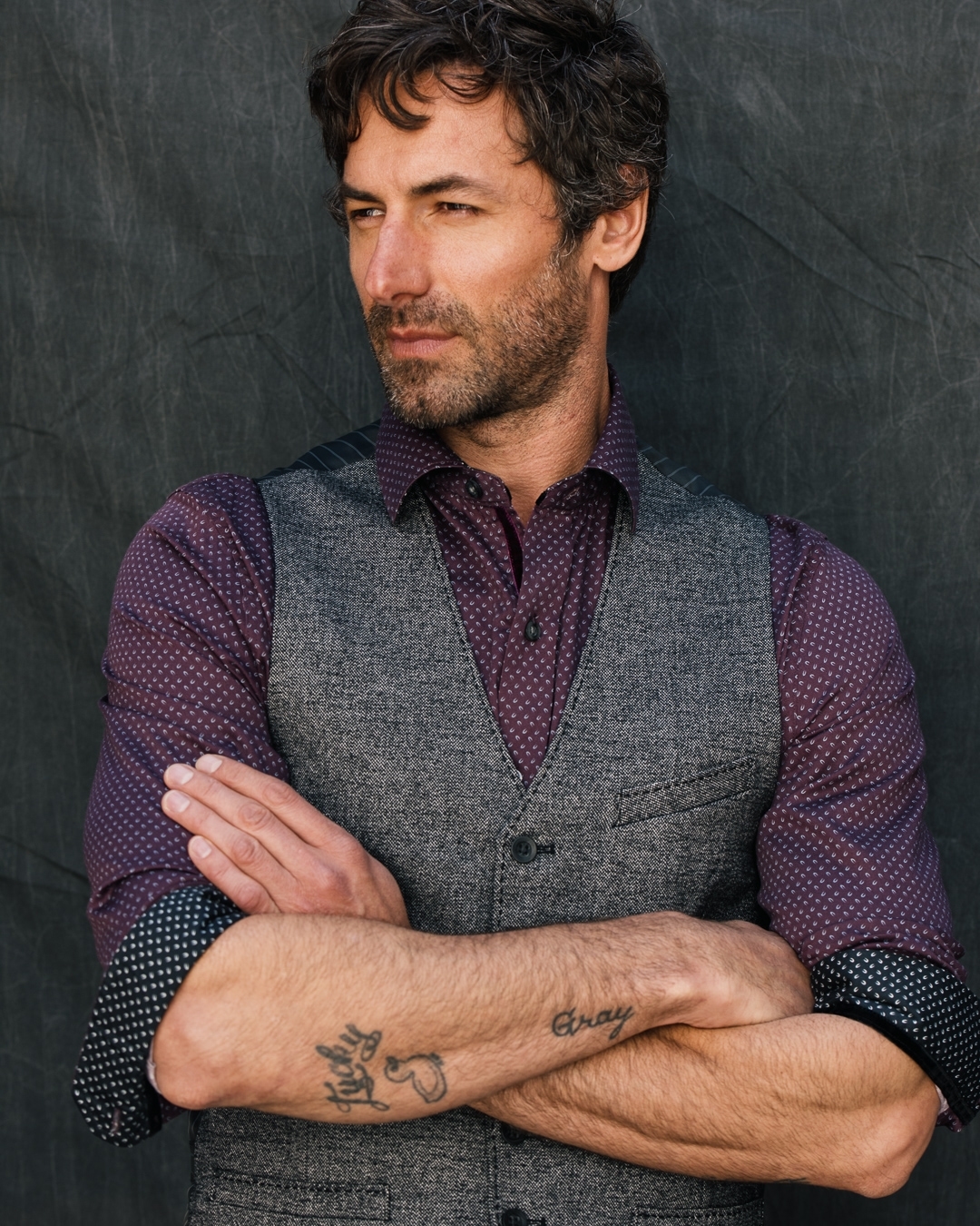Portrait of a man with dark hair and a vest in a photo studio, arms crossed and a patterned shirt. Gray backdrop.