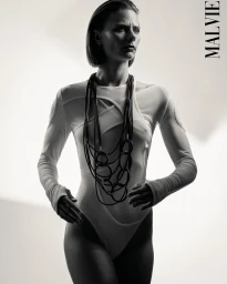 Model wearing a white bodysuit and statement black necklace against a light background, photographed by Geoffrey Badner