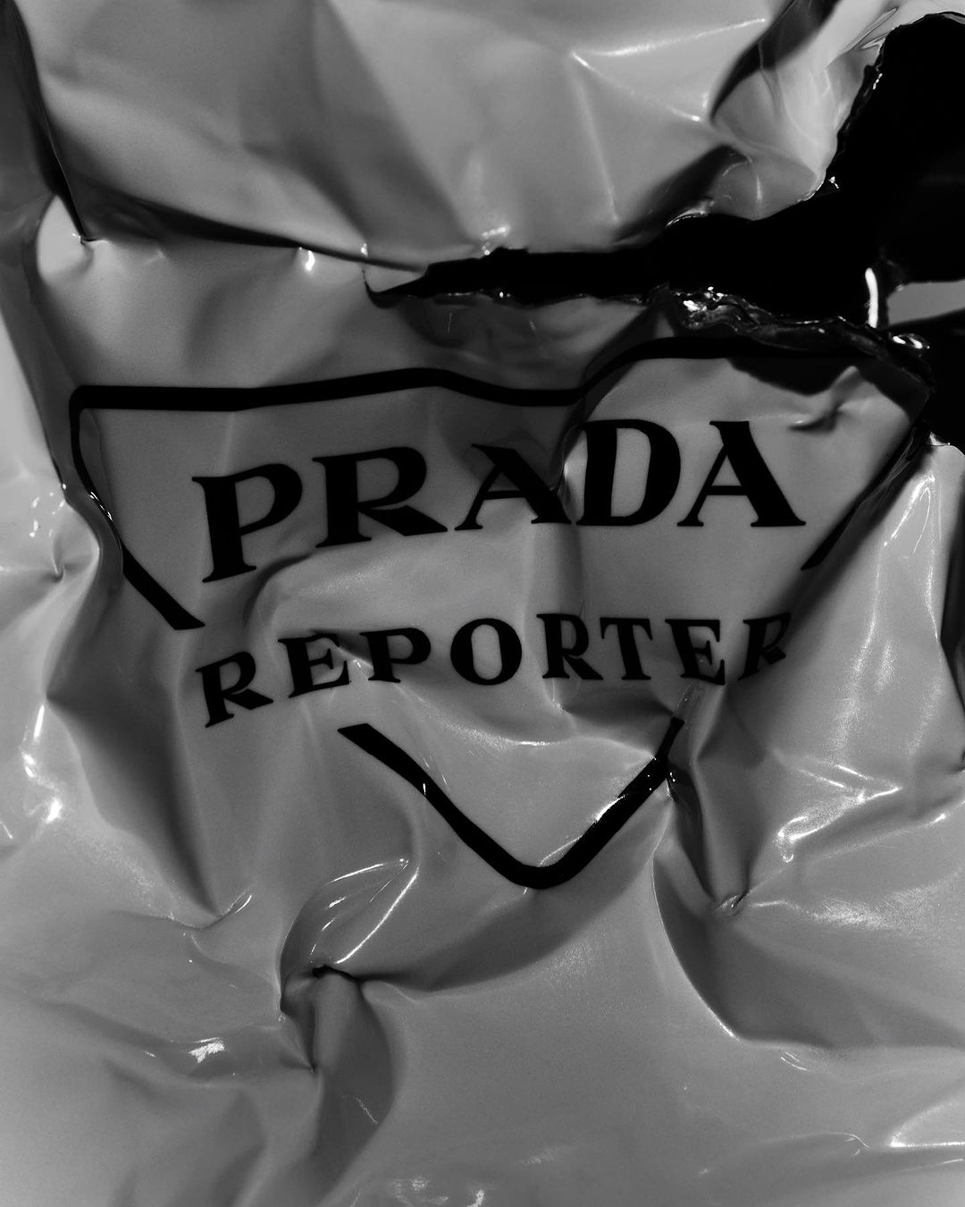 Close-up black and white shot of a crumpled glossy surface with the words 'PRADA REPORTER' prominently displayed.