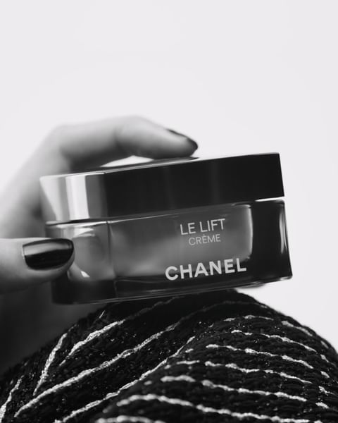 Close-up of a hand holding a jar of Chanel Le Lift Crème on a textured black and white surface, skincare product, beauty photography.