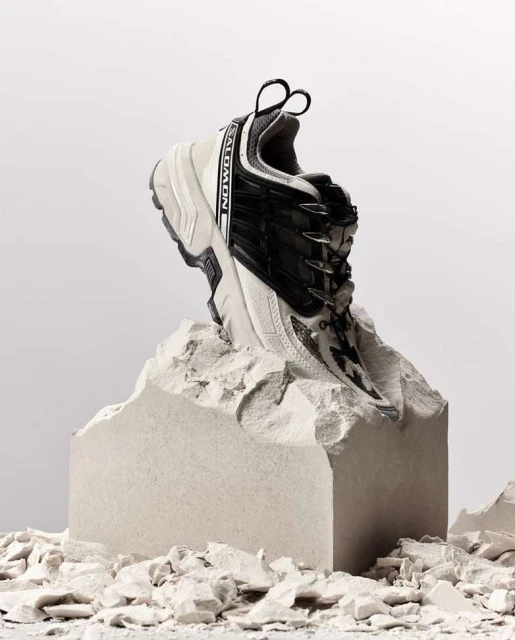 Black and white Salomon sneaker on a rough, light gray stone platform, studio lit against a light background. Fashion footwear.