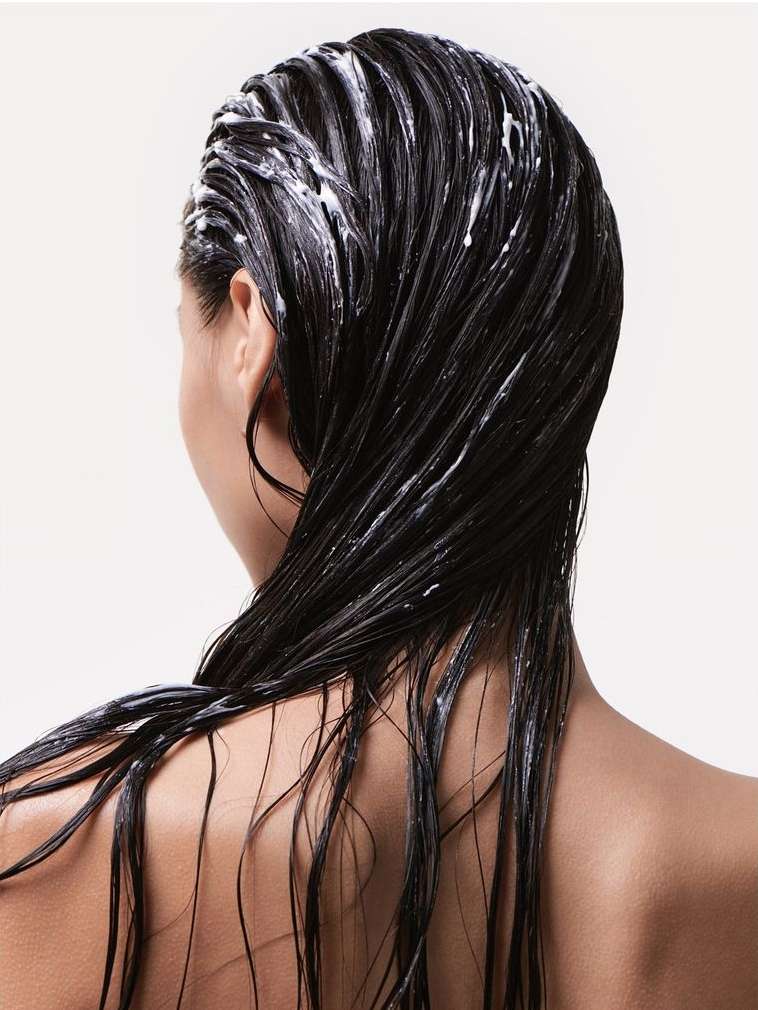 A woman's back and head are visible, showcasing dark, wet hair covered in white conditioner, set against a clean, light background.