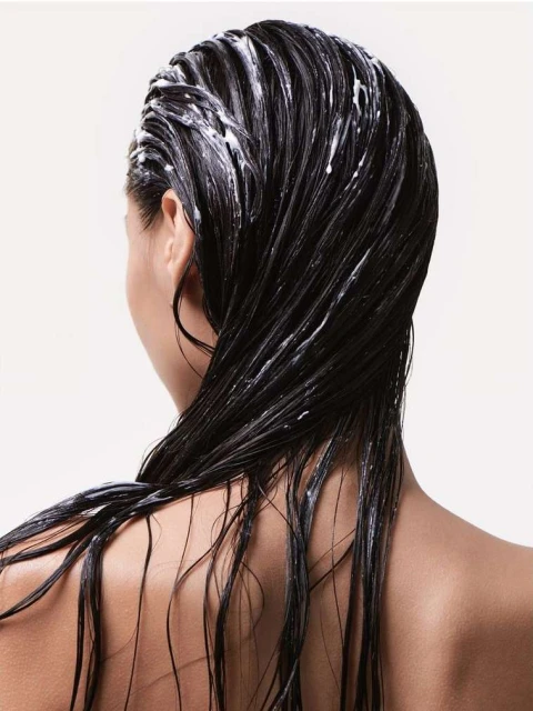 A woman's back and head are visible, showcasing dark, wet hair covered in white conditioner, set against a clean, light background.