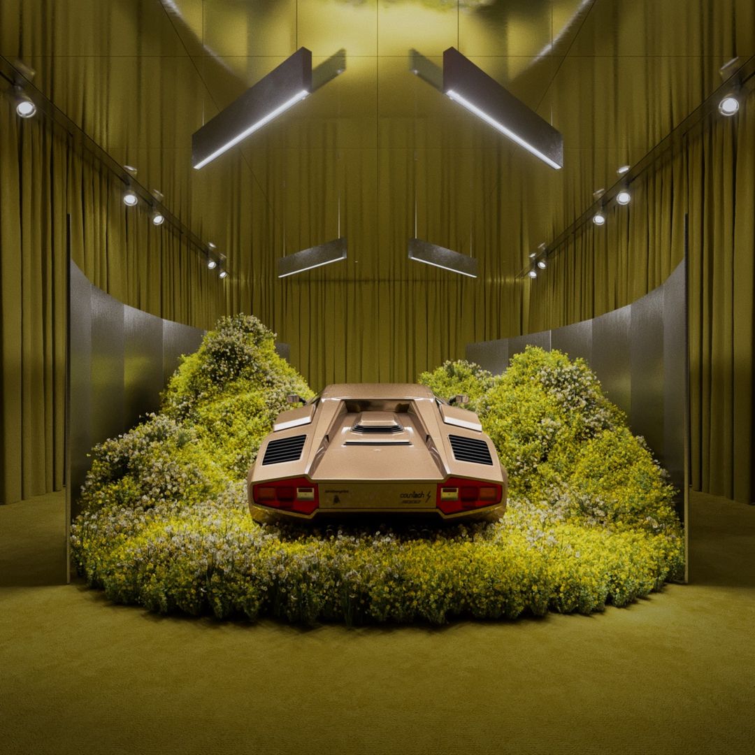 Rear view of a gold Lamborghini Countach displayed on a round platform covered in greenery, set against a yellow curtain backdrop.