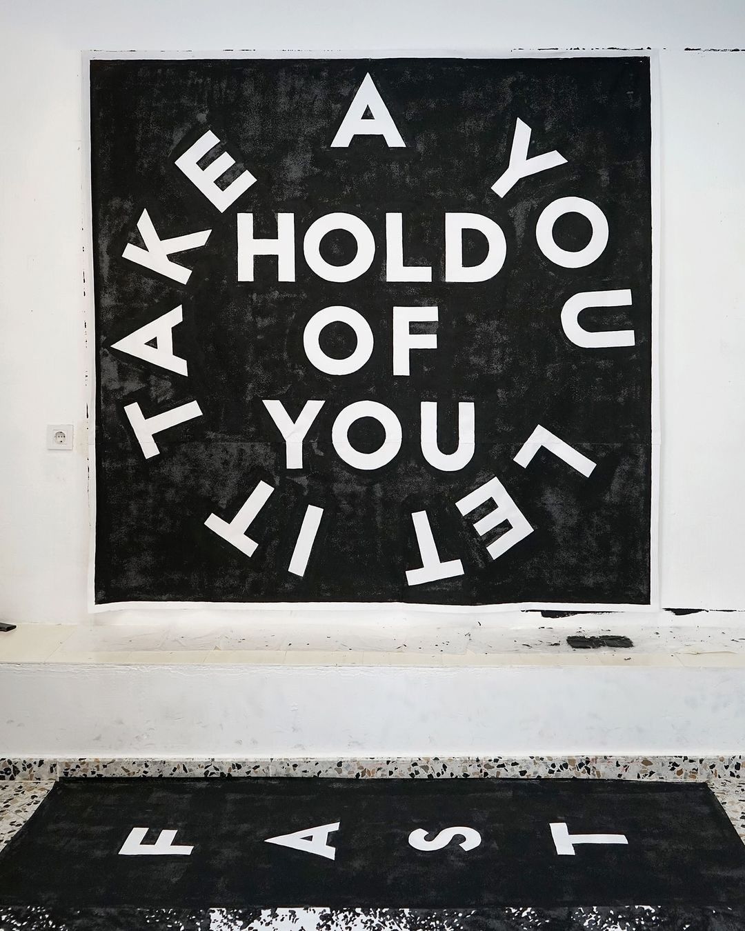 Artwork featuring a circular arrangement of white block letters on a black background, stating 'Take a hold of you let it fast'.