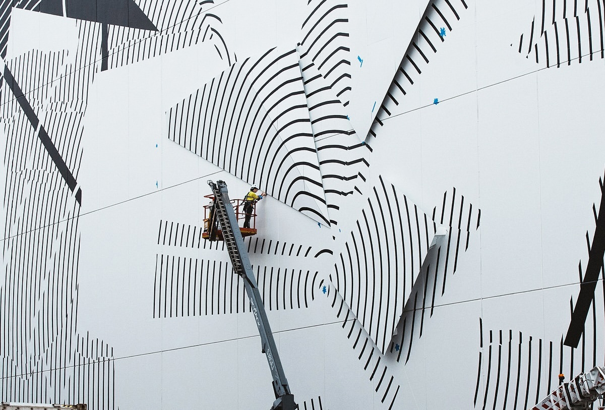 A worker on a mechanical lift repairs graphic art on the exterior wall of a building, featuring black lines on a white background.
