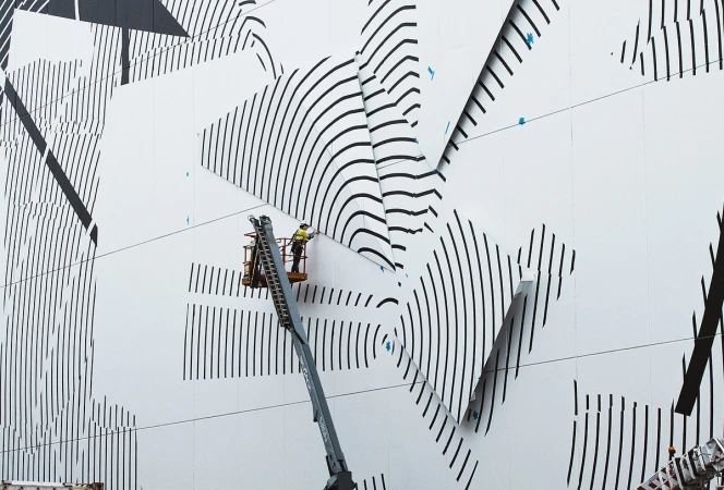 A worker on a mechanical lift repairs graphic art on the exterior wall of a building, featuring black lines on a white background.