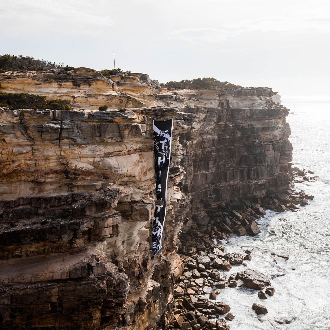 Coastal cliffside view featuring a black and white banner with text hanging down the rock face, set against a backdrop of the ocean.