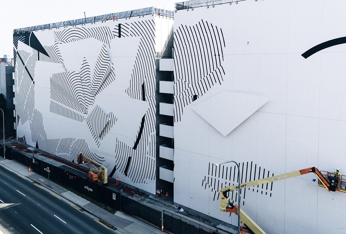 Exterior of a building featuring a black and white geometric mural, with construction and maintenance workers using a lift platform.