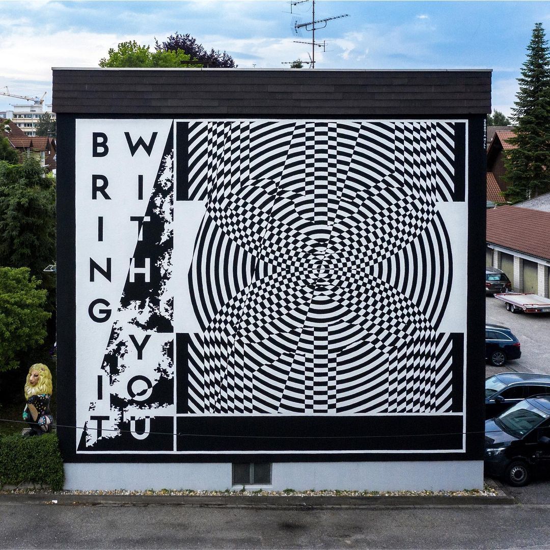 Eye-level shot of a building mural with a striking black and white geometric pattern, set against an urban backdrop with cars and foliage.