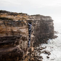 Large black typographic banner suspended from the edge of a rugged coastal sandstone cliff overlooking the ocean.