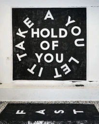 Large black square artwork with bold white lettering reading TAKE A HOLD OF YOU LET IT FAST mounted on a white wall above a patterned floor