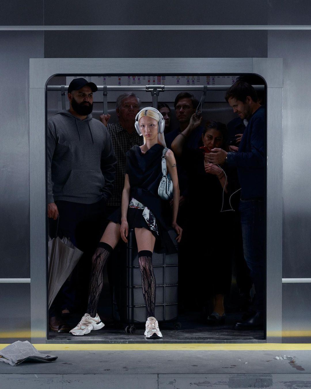 Fashion model in black dress and patterned tights stands out in a crowded subway elevator, accessorized with a purse and headphones.