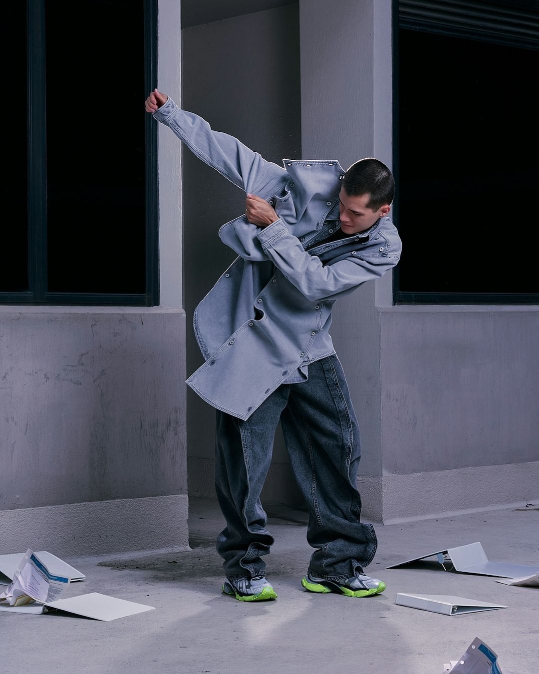 A male fashion model strikes a dynamic pose wearing a light blue denim jacket and dark jeans in an urban environment.