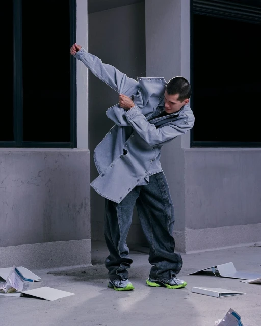 A male fashion model strikes a dynamic pose wearing a light blue denim jacket and dark jeans in an urban environment.