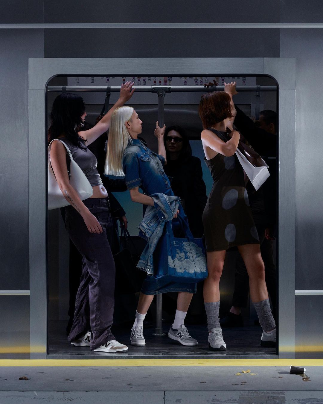 Three women in an underground train wearing denim and black dresses. The underground train is grey with a yellow line on the floor.