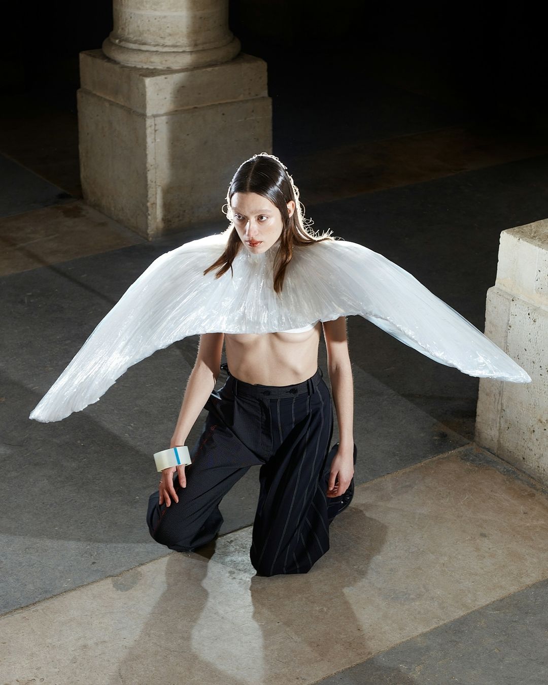 Woman poses in high-fashion with a translucent wing-like garment, on her knees in a stone court under dark conditions and bright highlights.