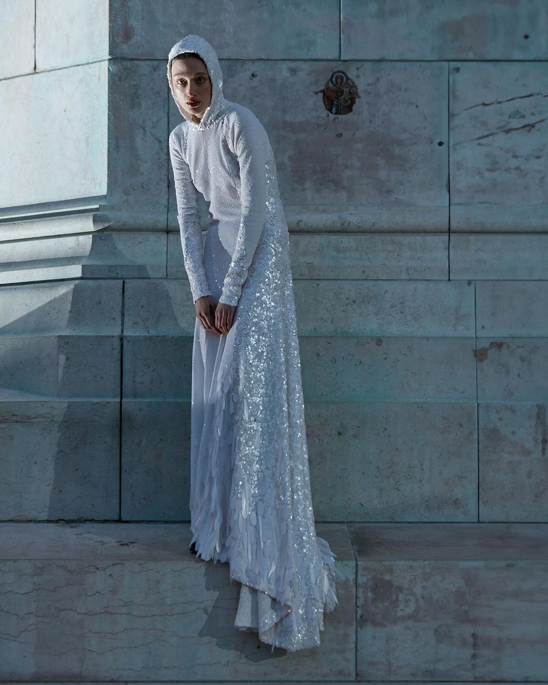 Adult female fashion model poses in a hooded, long-sleeved, floor-length white sequin dress on a light marble stone platform.