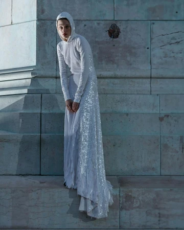 Adult female fashion model poses in a hooded, long-sleeved, floor-length white sequin dress on a light marble stone platform.