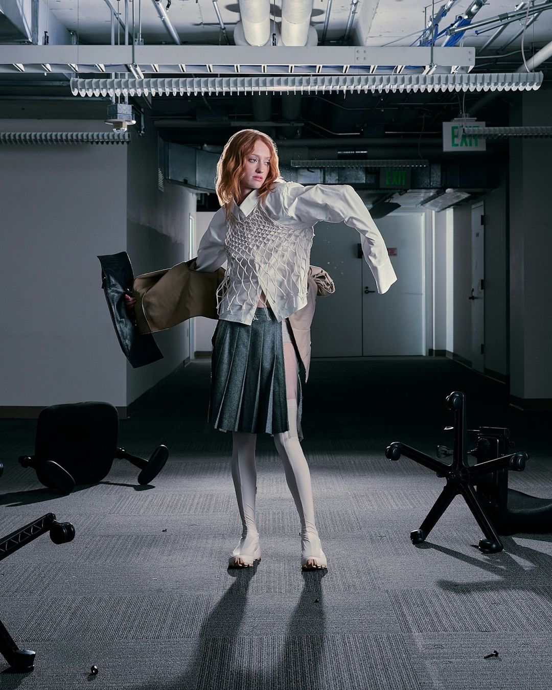 Fashionable woman with auburn hair in a deconstructed white shirt, textured vest, and pleated skirt poses among fallen chairs in an office.