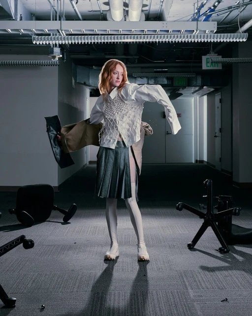 Fashionable woman with auburn hair in a deconstructed white shirt, textured vest, and pleated skirt poses among fallen chairs in an office.