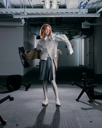 Fashionable woman with auburn hair in a deconstructed white shirt, textured vest, and pleated skirt poses among fallen chairs in an office.