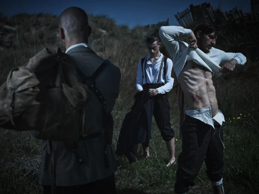 Three men near a beach at night. One man removes his shirt, revealing a scar on his abdomen. Another wears suspenders.