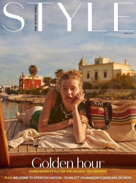 Magazine cover featuring a woman reclining on a boat, showcasing Loewe tote bag, set against a coastal backdrop with clear blue sky.