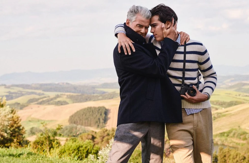 Two men embrace in a sunny outdoor setting. The older man wears a dark coat, while the younger man has a striped sweater and camera.