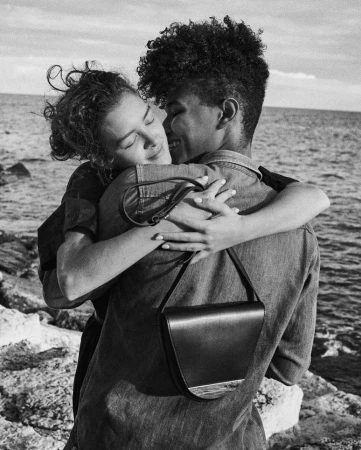 Two people embracing on a rocky beach with the sea and sky in the background. One person has a black purse hanging on their shoulder.