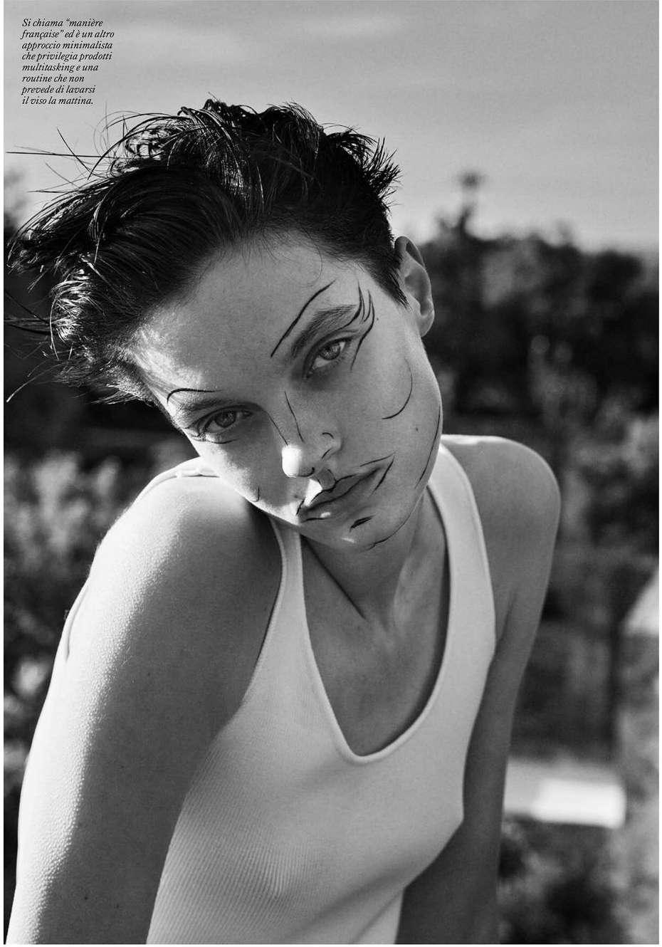 Monochrome portrait of a woman with short hair and artistic makeup looking at the camera outdoors against a natural background.
