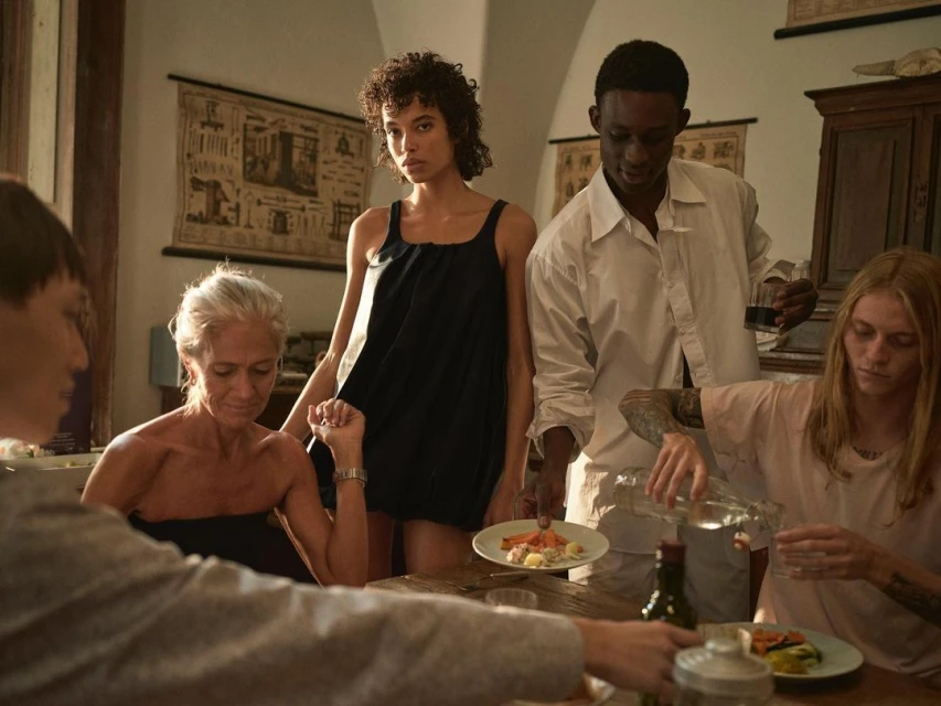 A group of diverse individuals gather around a table in a softly lit interior setting, sharing a meal with food and drinks visible.