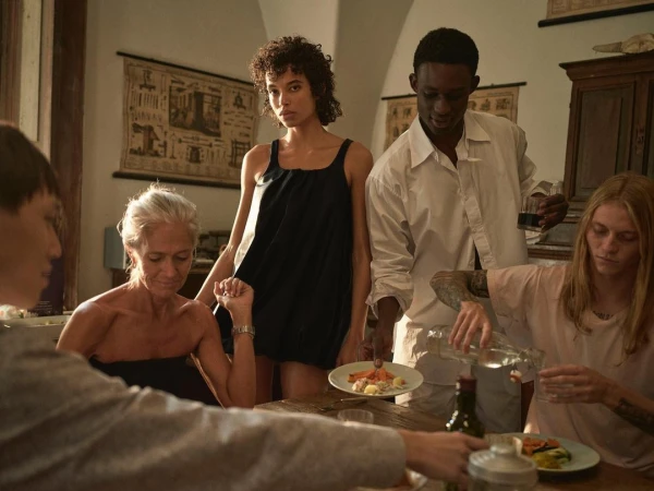 A group of diverse individuals gather around a table in a softly lit interior setting, sharing a meal with food and drinks visible.