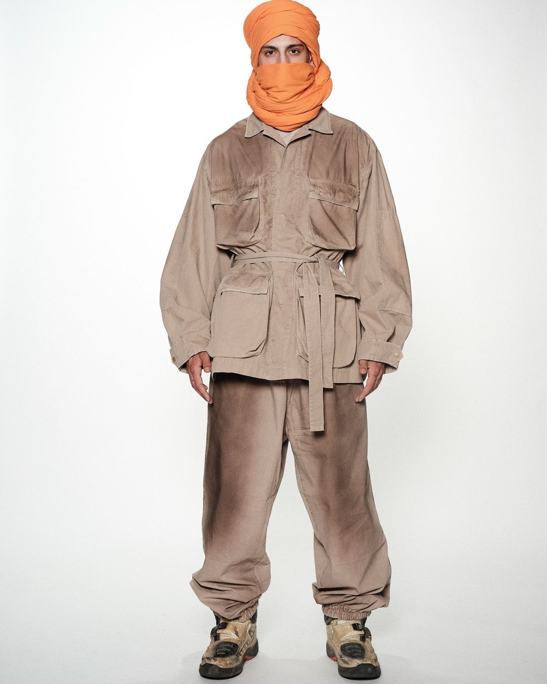 A man in a tan jacket, pants, and a wrapped orange scarf stands on a white background, looking at the camera with a neutral expression.