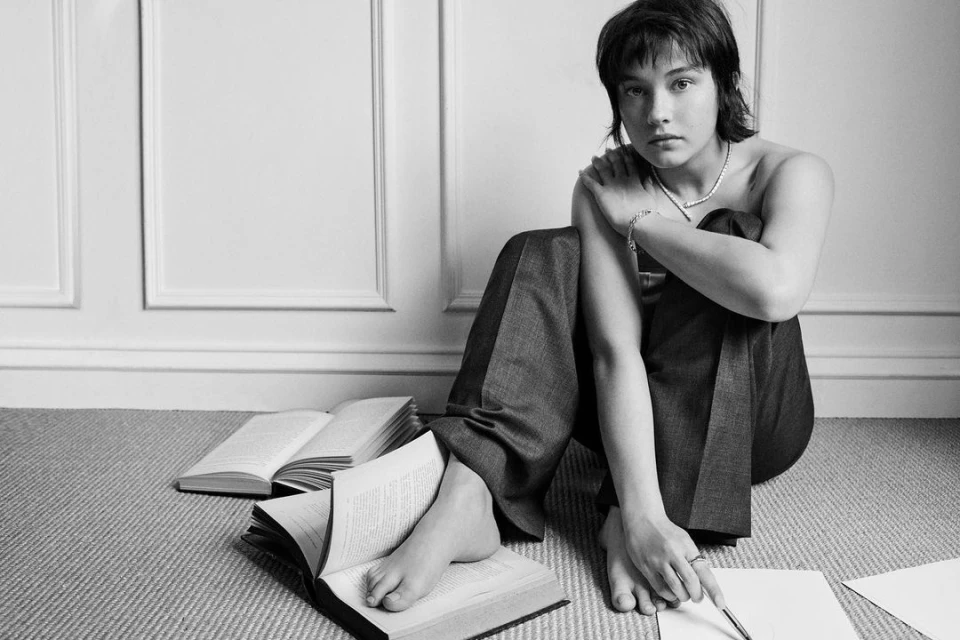 A young adult sits on the floor with open books and papers in a black and white photo, styled with a necklace and casual pants.