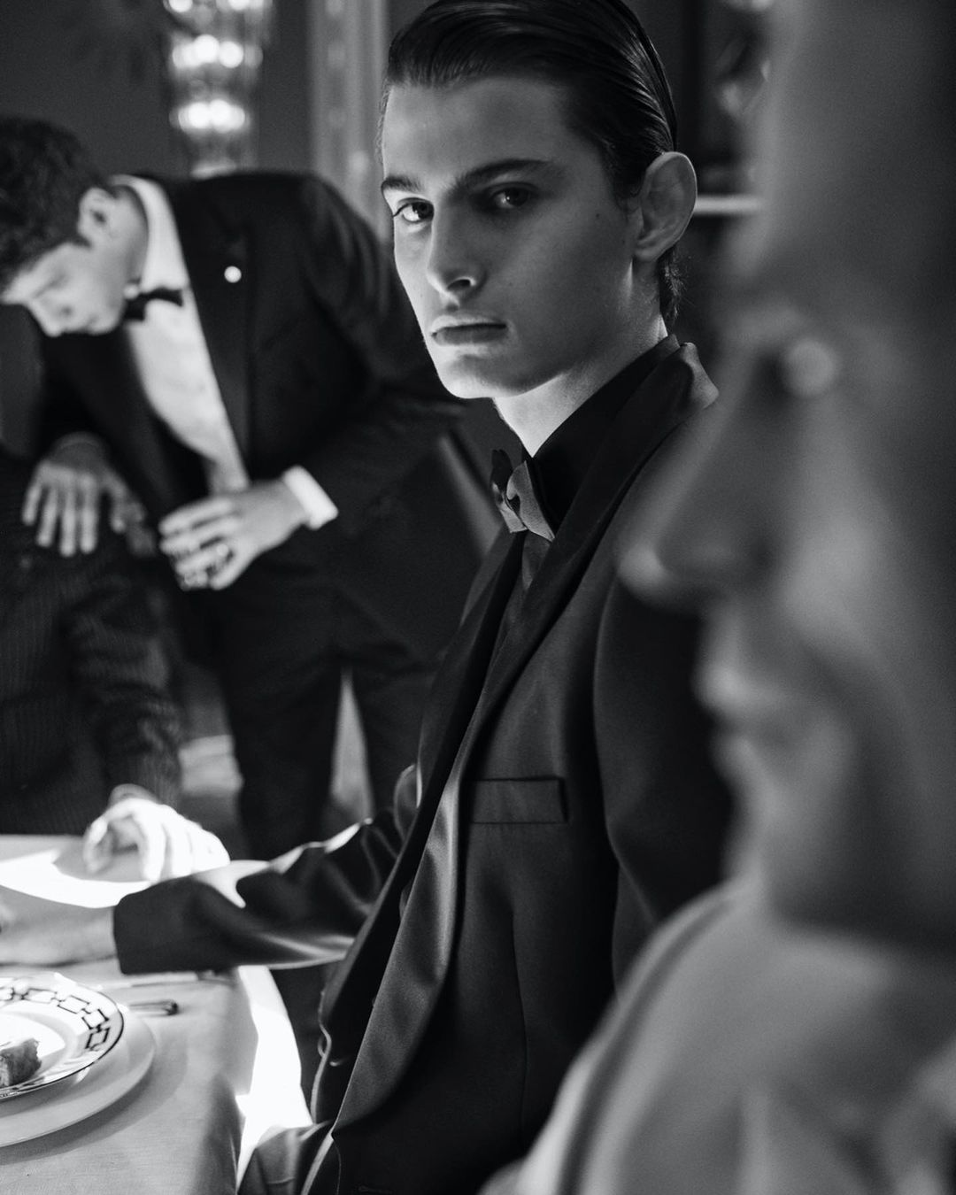 Black and white portrait of a young man in a black tuxedo, seated at a table with other men in tuxedos in the background.