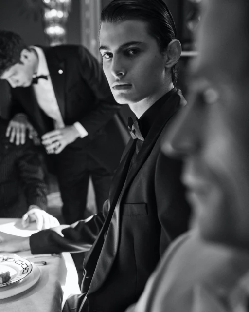 Black and white portrait of a young man in a black tuxedo, seated at a table with other men in tuxedos in the background.