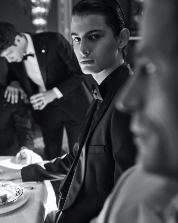 Black and white portrait of a young man in a black tuxedo, seated at a table with other men in tuxedos in the background.