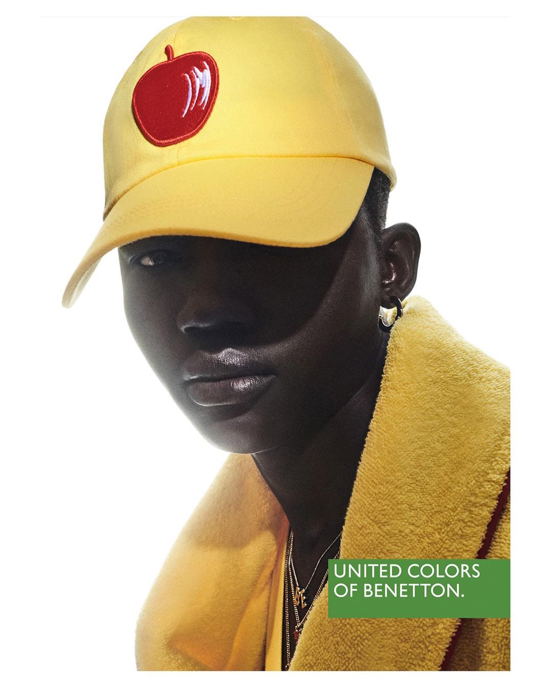 Dark-skinned person in a yellow cap with an apple emblem and a yellow jacket, posing indoors under bright studio lighting. Fashion shot.