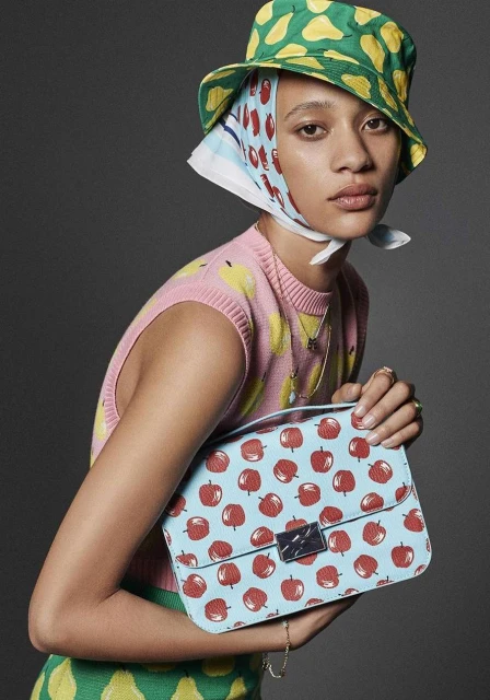Young woman poses in a pink sweater, bucket hat, and headscarf, holding a blue purse with red apples in a gray studio setting.