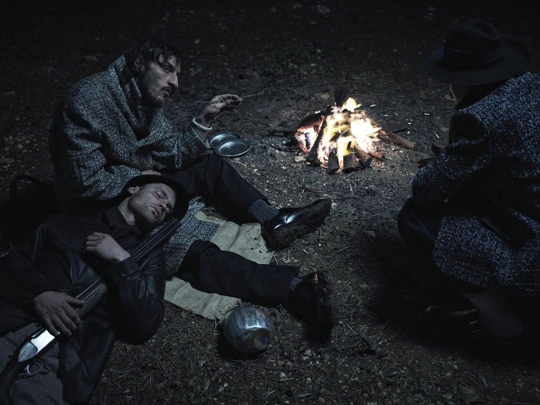 Three men gather around a campfire at night, one is sleeping with a rifle nearby. The scene is dimly lit, giving it a cinematic quality.