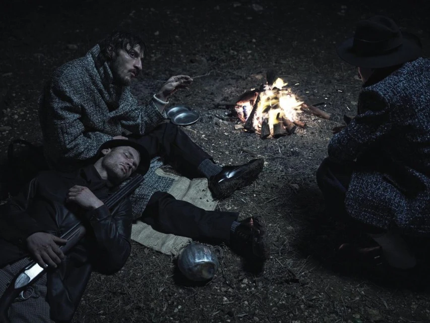 Three men gather around a campfire at night, one is sleeping with a rifle nearby. The scene is dimly lit, giving it a cinematic quality.
