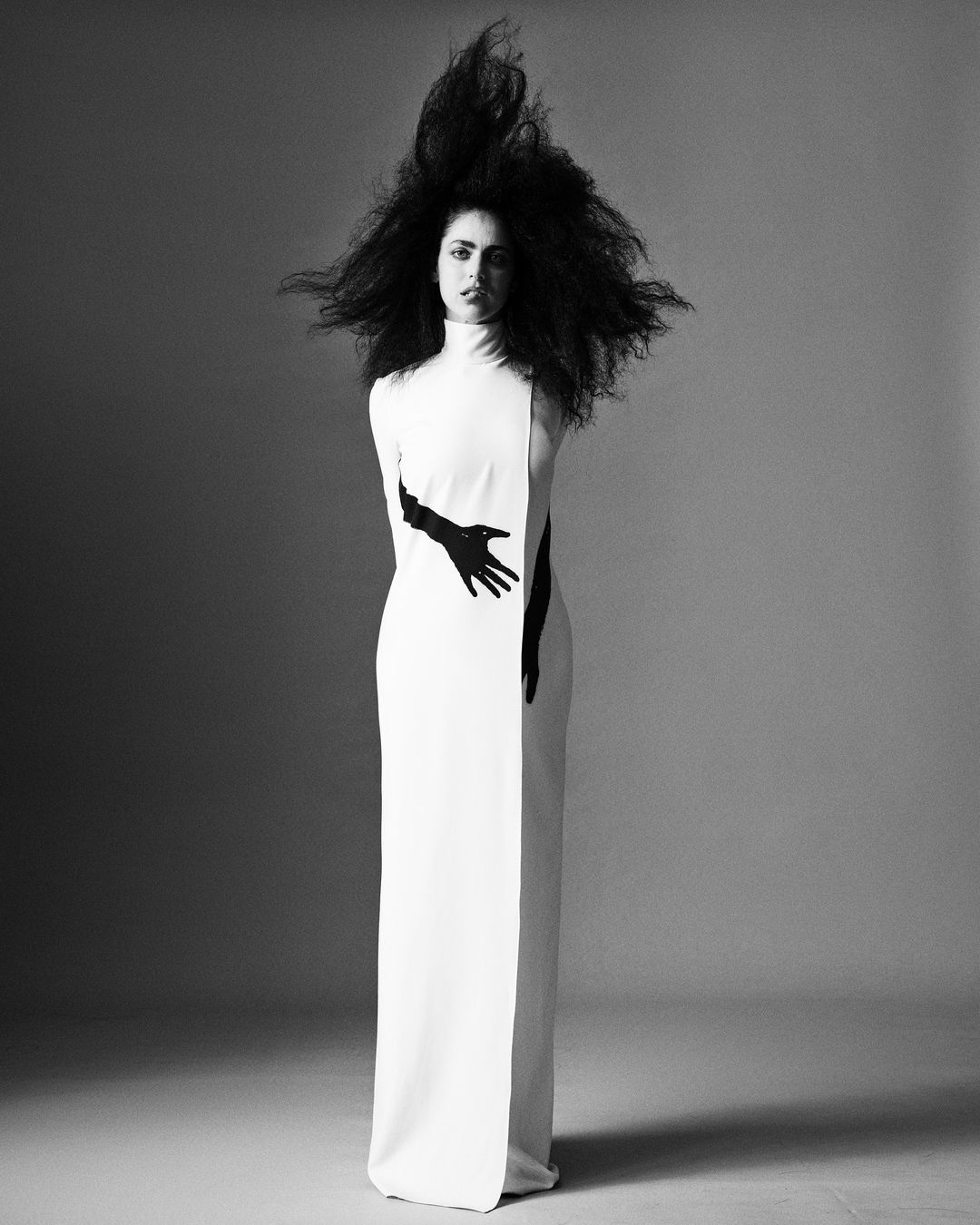 Monochrome portrait of a woman with voluminous dark hair wearing a white, high-necked gown with long black gloves in a studio setting.