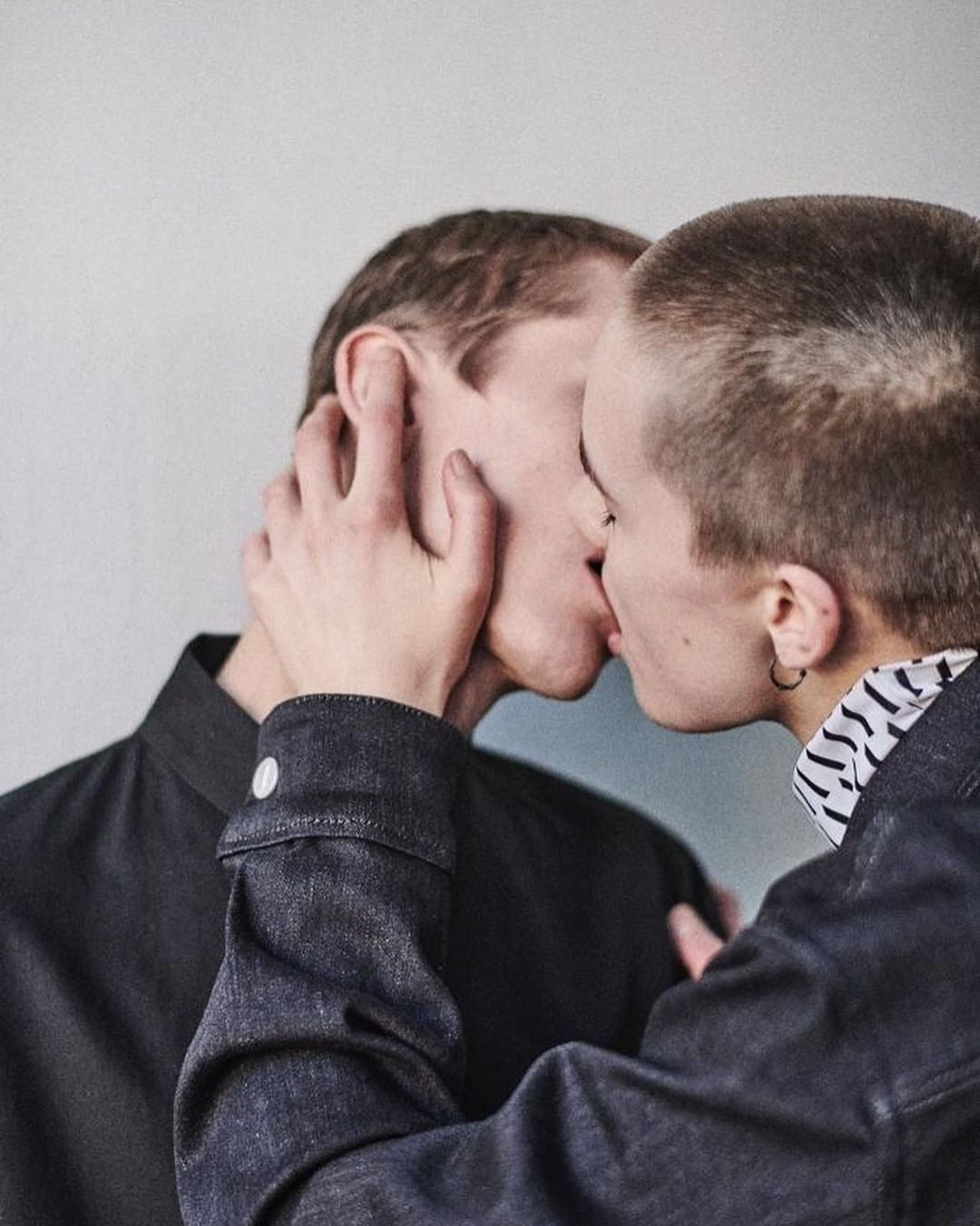 Close-up portrait of two young adult fair-skinned people kissing, wearing a dark shirt and jean jacket against a grey background.